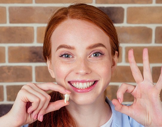 A smiling woman holding a removed tooth and making an ‘Okay’ sign with her hand