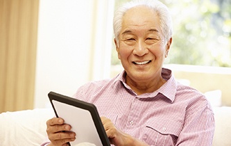 An older man seated on the couch and using a tablet while showing off his dental implants