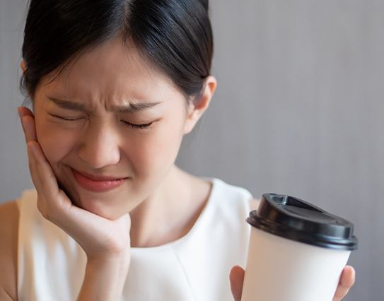 A young woman holding a coffee cup in one hand and her cheek in the other, cringing in pain because of sensitivity to her hot beverage